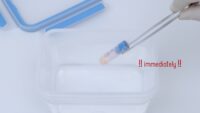 Forceps holding cryopreservation tube with blue cap over container with sterile water for thawing.