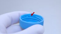 Blue lid of 50 mL centrifuge tube held in hand with gloves, red arrow indicating small droplets of liquid inside the thread of the lid.