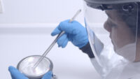 Handling samples in liquid nitrogen in a dewar with large forceps, wearing blue cryoprotective gloves, white lab coat and protective face shield.