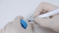 Person wearing disinfected white gloves labelling 50 mL centrifuge tube with blue cap with a lab marker.