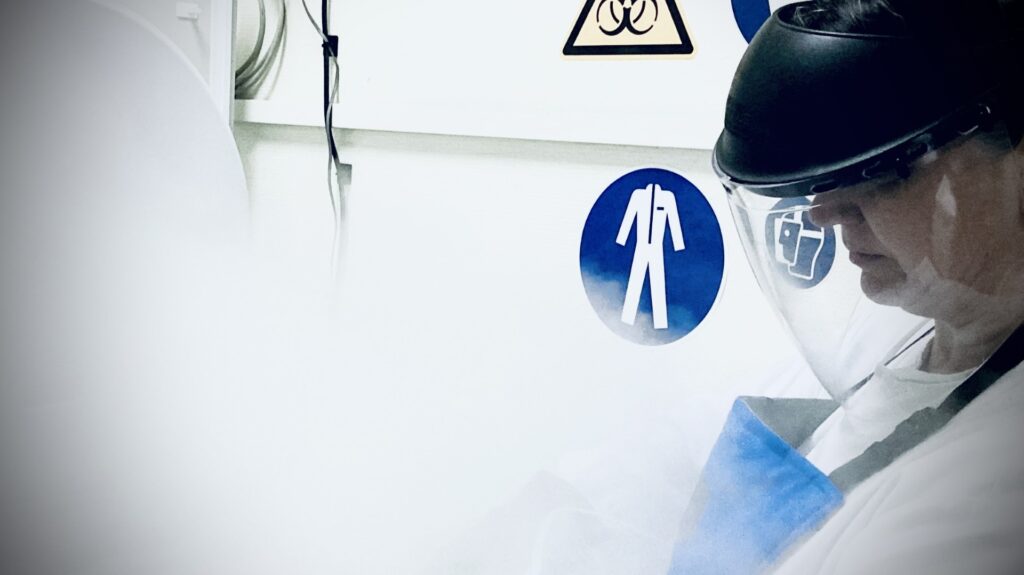 A person in white lab coat, face shield, and heavy cryoprotective apron in front of a liquid nitrogen cell storage tank and vapour condensing.