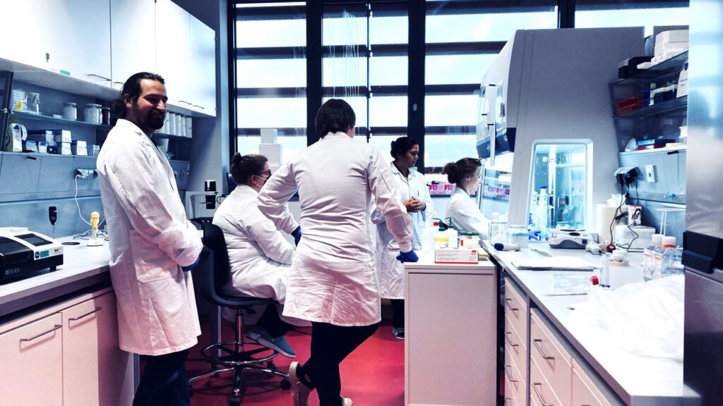 Five people in white lab coats in front of a laminar hood in a cell culture lab listening to practical instructions.