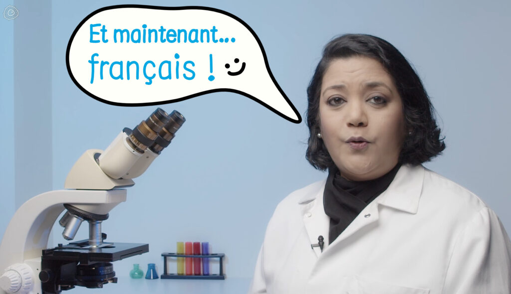 Women in white lab coat beside a laboratory microscope saying "Et maintenant.. Francais!"