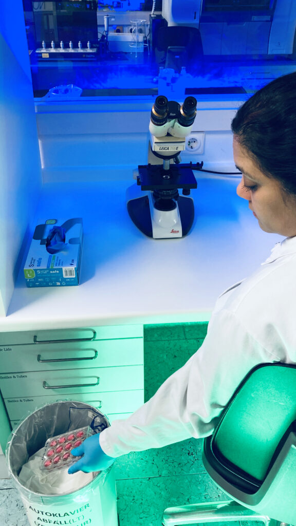 A person in the lab, wearing white lab coat and blue gloves, trashing a pile of cell culture plates into the autoclave waste bin.